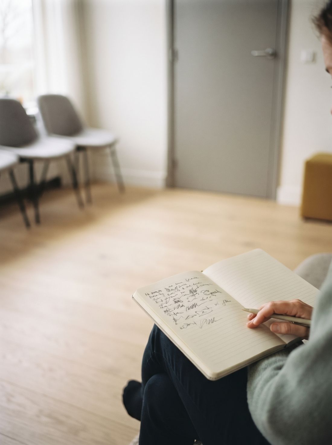 Un cuaderno abierto con anotaciones a mano, sobre las rodillas de un adulto en una sala de espera de una clínica.